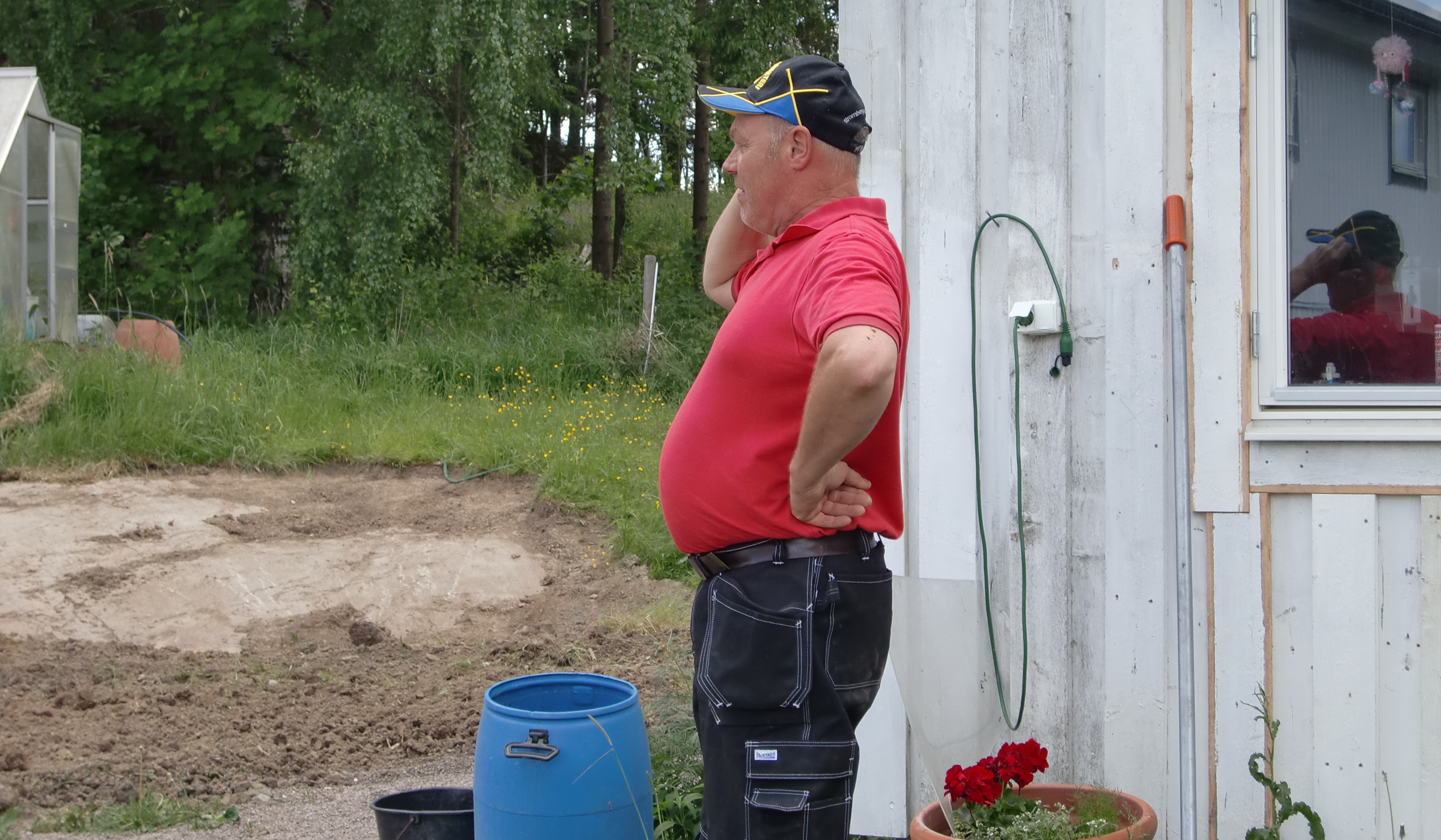 En bonde står utanför ett vitt hus och pratar i mobiltelefon.